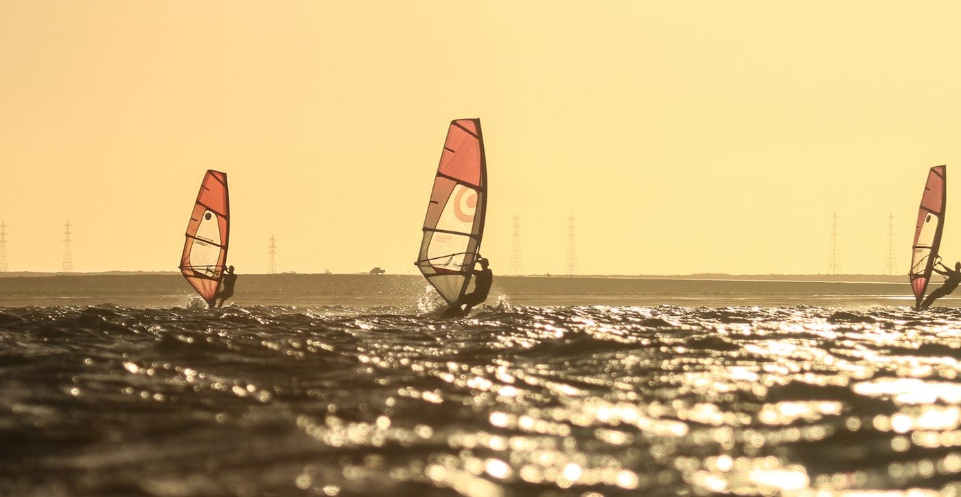Three windsurfers sailing across Dakhla Lagoon against a golden sky